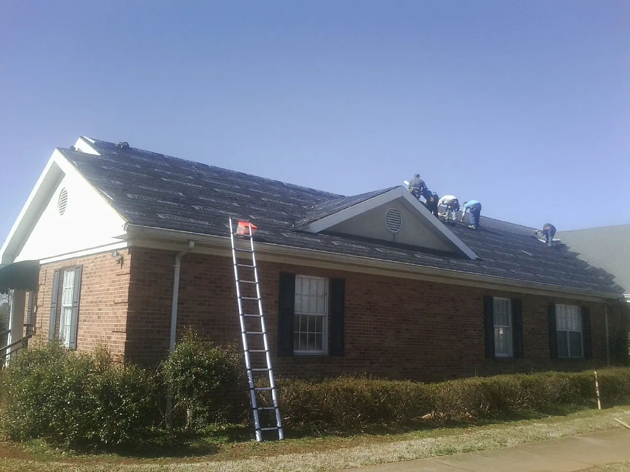 Crew tearing shingles off a brick ranch roof with a ladder set — Emergency Roof Repair work by our crew in Delanson