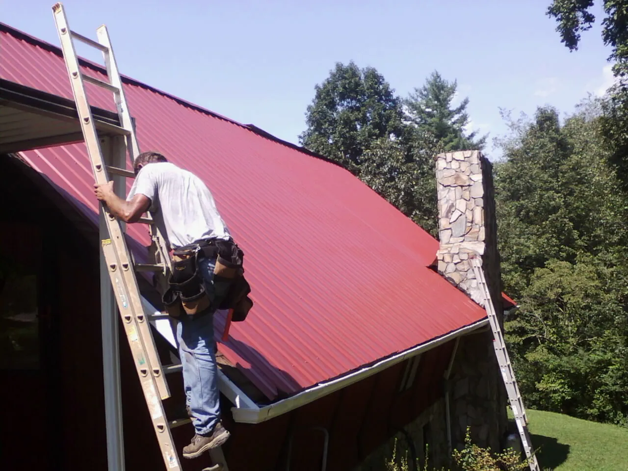Roofer on a ladder beside a red metal roof and stone chimney — Soffit & Fascia Repair by Roofing Brothers in Delanson